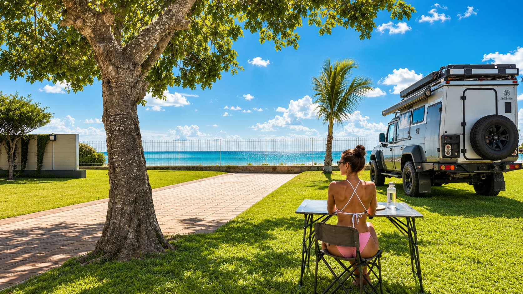 Guest relaxing at a camp table beside a 4x4 camper, shaded lawn overlooking the beach and ocean